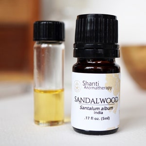 Op de afbeelding: Een klein amberkleurig glazen flesje met een zwarte dop gevuld met sandelhout etherische olie. Het etiket vermeldt "Shanti Aromatherapy SANDALWOOD Santalum album India .17 fl oz. (5ml)"