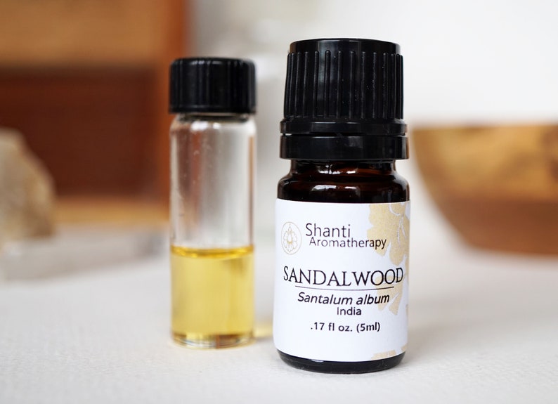 Op de afbeelding: Een klein amberkleurig glazen flesje met een zwarte dop gevuld met sandelhout etherische olie. Het etiket vermeldt "Shanti Aromatherapy SANDALWOOD Santalum album India .17 fl oz. (5ml)"
