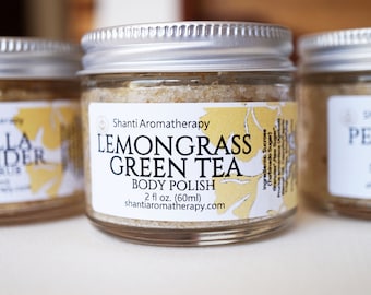 May include: Three glass jars with silver lids, labeled "Lemongrass Green Tea Body Polish," "Vanilla Lavender Sugar Scrub," and "Pepper Lemon Sugar Scrub." Each jar contains a grainy, light-colored product. The labels feature botanical designs and the brand name "Shanti Aromatherapy."