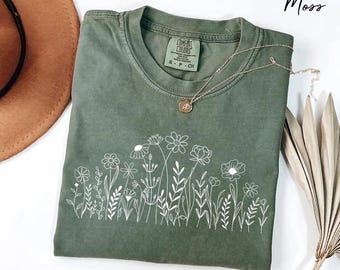May include: Olive green t-shirt with a white floral design across the front. The shirt has a crew neck and short sleeves. A gold necklace with a small pendant rests on the shirt. The word "Moss" is written in the upper right corner.