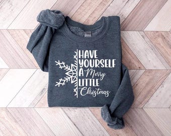 May include: A dark gray sweatshirt with the phrase "Have Yourself a Merry Little Christmas" in white script and a snowflake design. The sweatshirt is folded on a wooden surface.