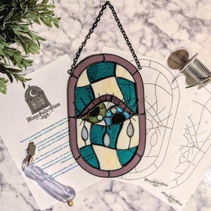 Op de afbeelding: Een ovaal glas-in-lood raamhanger met een centraal oogontwerp in teal, crème en paarse glassegmenten, verbonden door zwarte soldeerlijnen. Het object is voorzien van een zwarte metalen ophangketting en wordt getoond met papieren patronen.