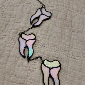 May include: A hanging decoration featuring three iridescent, tooth-shaped stained glass pieces connected by dark metal chains. Each tooth has a pearlescent finish with subtle pink and blue hues, outlined in dark solder, creating a vertical arrangement.
