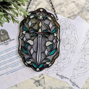 Può includere: Un acchiappasole in vetro colorato con un motivo centrale a spada circondato da intricati segmenti di vetro turchese, bianco e grigio uniti da linee di saldatura scure. L'oggetto include una catena nera per appenderlo e due modelli cartacei.