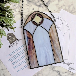 May include: Stained glass window panel with a black metal frame and chain. The panel features a light blue, brown, and purple color scheme with a geometric design. The image also includes design templates and a plant.