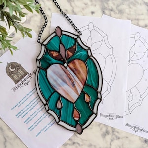 May include: A stained glass heart-shaped suncatcher with teal and brown glass, outlined in black. The suncatcher hangs from a black chain. Two pattern sheets and a plant are in the background.