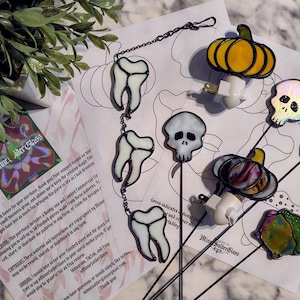May include: A collection of stained glass items including a three-tooth hanging chain, two plug-in night lights shaped like a yellow pumpkin and a multi-colored pumpkin, two skull-shaped plant stakes, a tooth stake, and a multi-colored abstract stake.