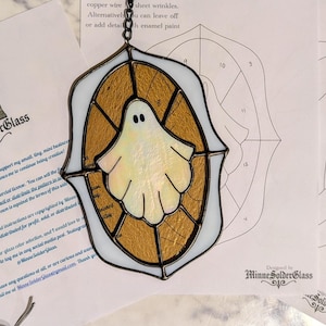 May include: A stained glass ghost suncatcher with a white body and gold-toned background, framed in dark metal solder. The piece is displayed over printed paper patterns featuring the MinneSolderGlass logo and ghost line art illustrations.