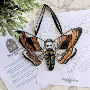 Peut inclure: Un ornement en vitrail en forme de papillon de nuit avec un motif de crâne central, des ailes en verre texturé ambre et blanc, et des lignes de soudure noires. L'objet inclut un ruban noir et repose sur des patrons en papier MinneSolderGlass.