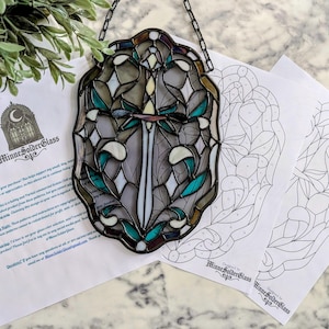 May include: A stained glass suncatcher featuring a central sword motif surrounded by teal and grey glass pieces in a scalloped frame, accompanied by printed paper patterns labeled MinneSolderGlass with black outlines and numbered sections.
