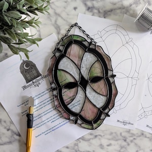 May include: Stained glass art piece with a black metal frame, featuring pink, white, and green glass panels. The piece is hanging from a chain. The image also shows a printed pattern, a glass cutter, and the text "MinneSolderGlass".