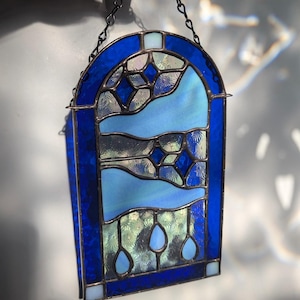May include: A stained glass window panel with a dark blue border and chain. The panel features shades of blue and clear glass, with a design of stylized water droplets and abstract shapes. The panel is approximately 6 inches wide and 8 inches tall.