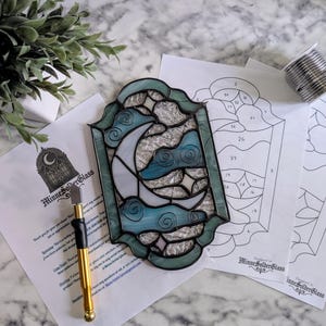 Op de afbeelding: Een glas-in-loodpaneel met een maan- en wolkenontwerp in blauw en wit, omlijst door een donkergroene rand. Inbegrepen zijn patroonvellen, een glassnijder en een spoel zilversoldeer. Het paneel is gemarkeerd met "MinneSolderGlass".