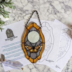 May include: Stained glass moth design in yellow and grey, with a chain for hanging. The artwork is displayed with pattern sheets and tools. The piece has a circular moon-like shape in the centre. The artwork is from MinneSolderGlass.