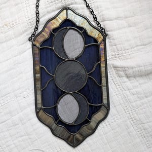 May include: Stained glass moon phases wall hanging. The piece features three circular moon phases in shades of gray and blue, set against a dark blue background. The frame is a dark metal with a decorative chain for hanging.