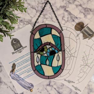 May include: Stained glass art piece featuring an eye design with teal, white, and black glass. The piece is framed in a purple border and hangs from a black chain. The image includes a glass cutter, wire, and pattern sheets.