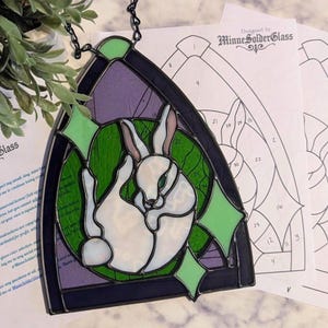 May include: A stained glass suncatcher featuring a white rabbit curled in a circle against a green circular background, framed by purple glass and light green diamond accents. The piece includes a black metal chain and printed pattern templates.