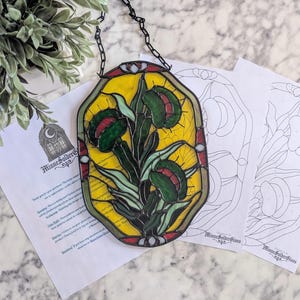 May include: A stained glass panel featuring a Venus flytrap design with yellow, green, and red colors. The panel is framed with a black chain for hanging. Two line art drawings are next to the panel.