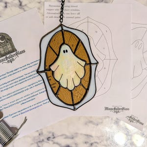 Op de afbeelding: Een glazen spookhanger met een wit en goud ontwerp, hangend aan een ketting. De hanger heeft een witte rand. De afbeelding bevat ook patroonvellen en een glassnijder. De tekst "MinneSolderGlass" is zichtbaar.