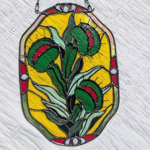 May include: Stained glass panel featuring a Venus flytrap design. The artwork showcases a yellow background with green and red flytraps, outlined in black. The panel is framed with red and white accents and is suspended by a black chain.