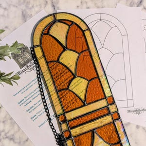 May include: A stained glass window panel with a chain, featuring a design of orange and yellow glass pieces. The panel is next to pattern templates. The text "MinneSolderGlass" is visible on the panel and templates.