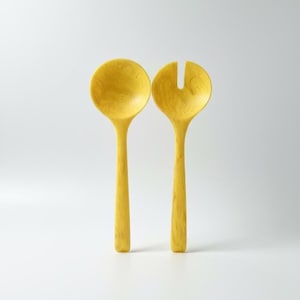 May include: A pair of yellow salad servers. One is a spoon, the other a fork. Both have long, slender handles and rounded heads. The utensils are made of a material with a marbled texture. The background is a plain white.