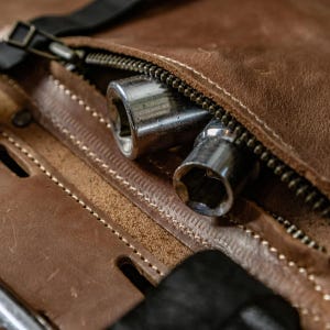 May include: Close-up of a brown leather tool bag with a silver zip. Two silver socket wrenches are partially visible inside the open bag. The bag has visible stitching and a black strap.