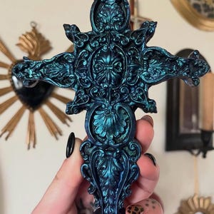 Puede incluir: Cruz adornada de color turquesa con intrincados detalles florales y de volutas. La cruz tiene un brillo metálico y un diseño simétrico detallado. La cruz se sostiene en una mano con esmalte de uñas negro.