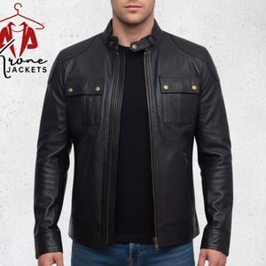 K&ouml;nnte beinhalten: Schwarze Lederjacke mit Stehkragen, zwei Brusttaschen mit goldfarbenen Knopfverschl&uuml;ssen und einem Frontrei&szlig;verschluss. Die Jacke wird &uuml;ber einem schwarzen Hemd und blauen Jeans getragen. Der Markenname "Krone Jackets" ist oben links sichtbar.