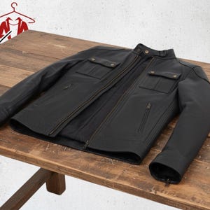 K&ouml;nnte beinhalten: Schwarze Lederjacke mit Rei&szlig;verschluss, zwei Brusttaschen und zwei Seitentaschen. Die Jacke liegt auf einem Holztisch. Sie hat einen Stehkragen und Rei&szlig;verschlussmanschetten.