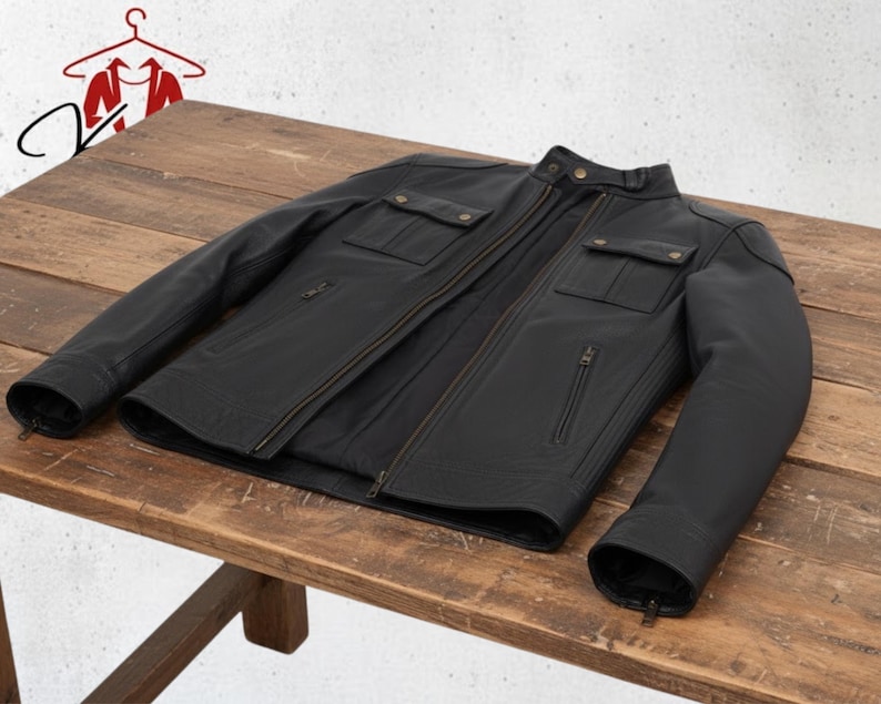 K&ouml;nnte beinhalten: Schwarze Lederjacke mit Rei&szlig;verschluss, zwei Brusttaschen und zwei Seitentaschen. Die Jacke liegt auf einem Holztisch. Sie hat einen Stehkragen und Rei&szlig;verschlussmanschetten.