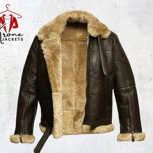 Puede incluir: Chaqueta de cuero marrón oscuro con forro y cuello de borrego beige. La chaqueta tiene cierre de cremallera, cinturón en la cintura y ribete de borrego en los puños. El logo "Krone Jackets" es visible.