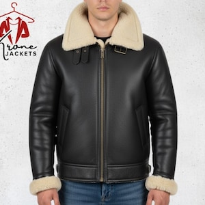 Puede incluir: Chaqueta de cuero negro con cuello y puños de borrego color crema. La chaqueta tiene cremallera, dos bolsillos y una correa con hebilla en el cuello. El logotipo de la marca "Krone Jackets" es visible en la esquina superior izquierda.