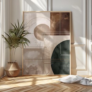 May include: Abstract art print in a gold frame. The artwork features geometric shapes and watercolor-style washes in shades of brown, beige, and dark green. Thin lines overlay the shapes. A potted plant and a white blanket are in the foreground.