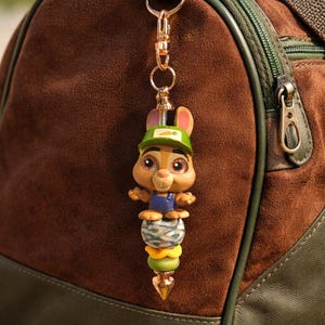 Op de afbeelding: Een goudkleurige sleutelhanger met een cartoonkonijn met een groene pet en blauwe overall. Het konijn zit bovenop een stapel kleurrijke kralen. De sleutelhanger is bevestigd aan een bruine sporttas met groene accenten.