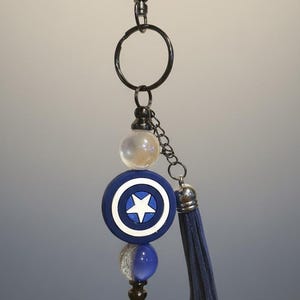 May include: A keychain featuring a blue and white shield design with a star in the center, a blue tassel, and various beads. The keychain is attached to a metal clasp.