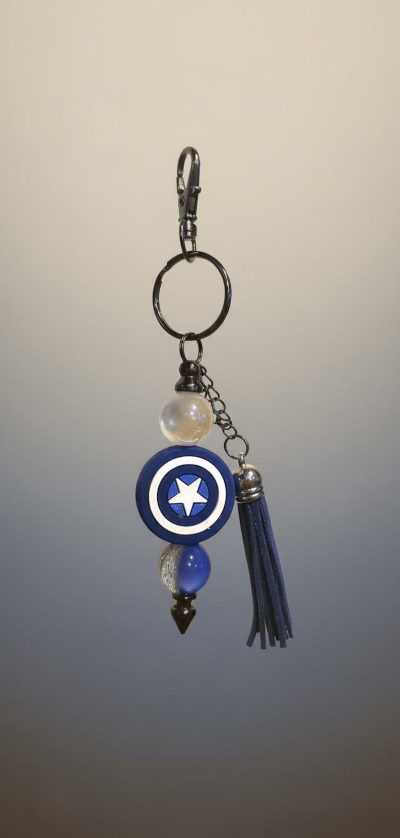 May include: A keychain featuring a blue and white shield design with a star in the center, a blue tassel, and various beads. The keychain is attached to a metal clasp.