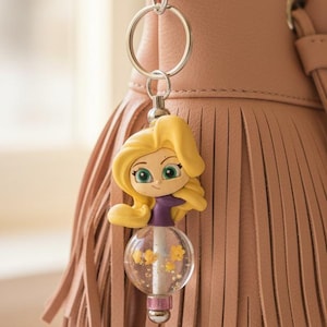 May include: A Rapunzel-themed keychain with a silver clasp, hanging from a pink fringed purse. The keychain features a cartoon figure with blonde hair, green eyes, and a purple top, holding a clear orb with yellow star accents.