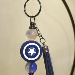 May include: A Captain America shield keychain with a blue, white, and silver color scheme. The keychain features a blue tassel and a black metal ring and chain.