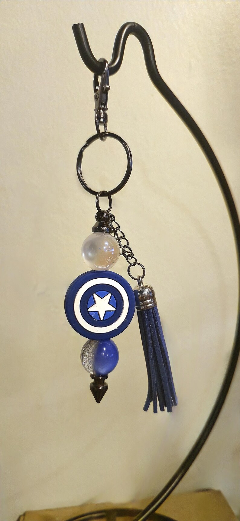 May include: A Captain America shield keychain with a blue, white, and silver color scheme. The keychain features a blue tassel and a black metal ring and chain.