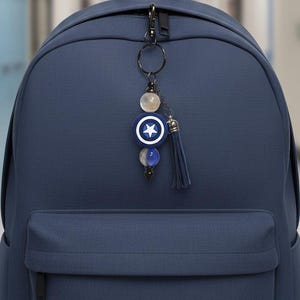 May include: A navy blue backpack with a decorative keychain. The keychain features a blue and white star emblem, beads, and a navy tassel. The backpack has a front pocket and a top handle.