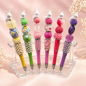 Könnte beinhalten: Eine Sammlung dekorativer Stifte mit verschiedenen farbenfrohen Perlenmustern. Jeder Stift hat einen weißen Hasenaufsatz und wird in einem transparenten Acrylhalter präsentiert. Die Stifte haben gelbe, grüne, rosa und violette Schäfte mit aufwendigen Perlenarbeiten.
