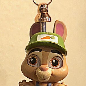 Op de afbeelding: Een grillige sleutelhanger met een cartoon konijn karakter met een groene pet met een wortel embleem. Het konijn draagt een blauwe overall en staat bovenop een decoratieve, kleurrijke kralen stapel. De sleutelhanger heeft een goudkleurige sluiting.