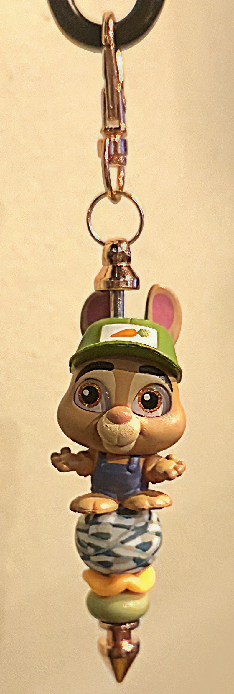 Op de afbeelding: Een grillige sleutelhanger met een cartoon konijn karakter met een groene pet met een wortel embleem. Het konijn draagt een blauwe overall en staat bovenop een decoratieve, kleurrijke kralen stapel. De sleutelhanger heeft een goudkleurige sluiting.