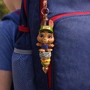 Op de afbeelding: Een grillige konijnen sleutelhanger met een groene pet en wortel detail, bevestigd aan een blauwe rugzak. De sleutelhanger heeft een stapel kleurrijke kralen en een goudkleurige sluiting, wat een speelse touch aan de rugzak toevoegt.