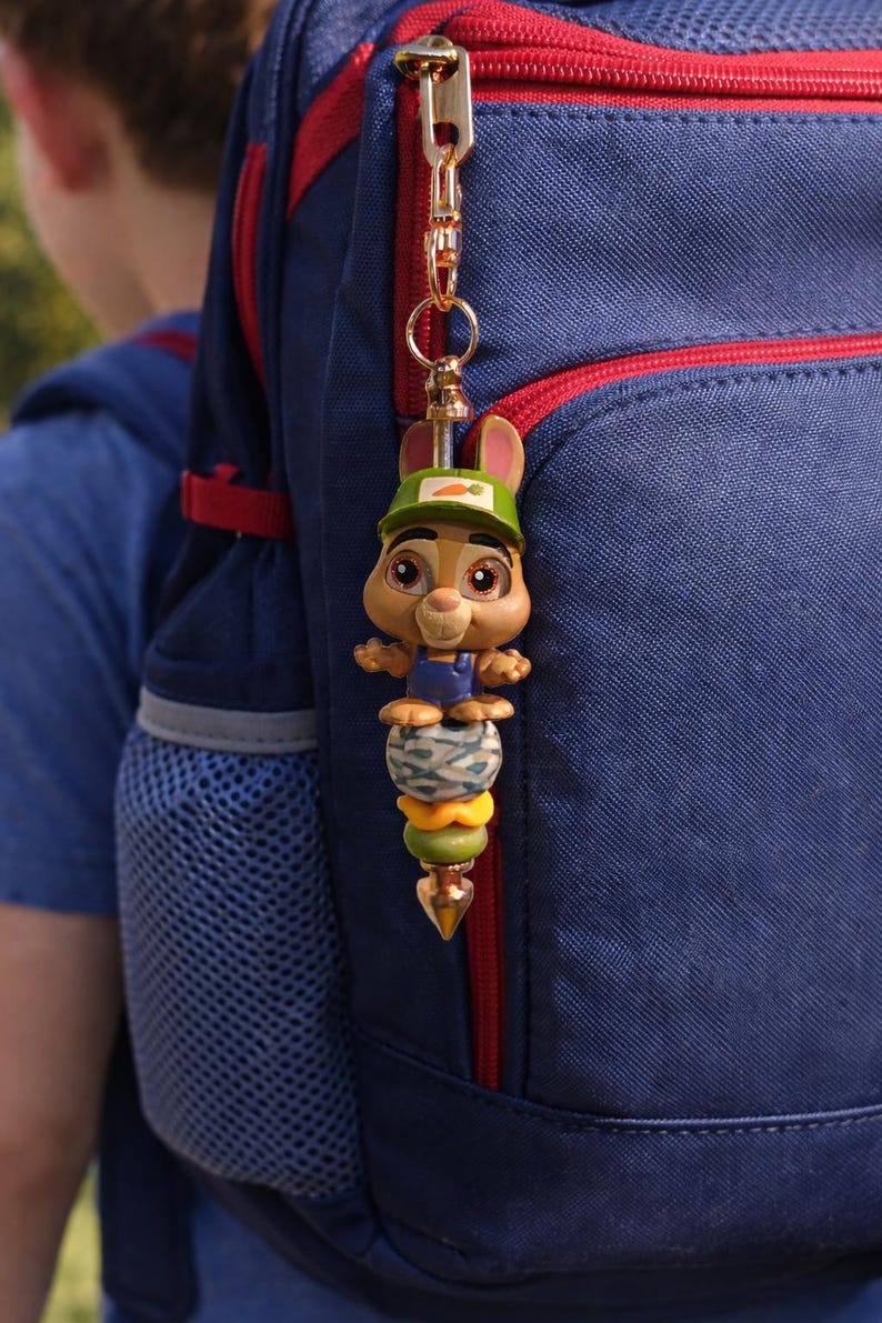 Op de afbeelding: Een grillige konijnen sleutelhanger met een groene pet en wortel detail, bevestigd aan een blauwe rugzak. De sleutelhanger heeft een stapel kleurrijke kralen en een goudkleurige sluiting, wat een speelse touch aan de rugzak toevoegt.