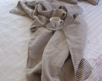 May include: A neutral-toned bedroom scene featuring a bed with white linens and two pillows. A folded blanket with striped detailing and a small teacup and saucer are placed on the bed, creating a cozy and inviting atmosphere.