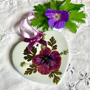 May include: A heart-shaped, white painted ornament decorated with a pressed purple flower and dried leaves. A purple ribbon and bow are attached for hanging. A purple flower and green leaves are placed above the ornament.