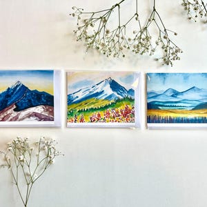 May include: Three watercolor paintings of mountain landscapes. Each painting features a different mountain scene with varying colors of blue, green, yellow, and red. The paintings are displayed in clear plastic sleeves.