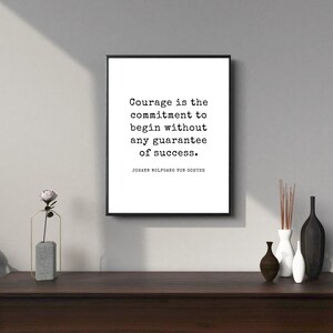 May include: A framed print with the quote "Courage is the commitment to begin without any guarantee of success" by Johann Wolfgang von Goethe. The print is displayed above a dark wooden cabinet with decorative vases and a small plant.